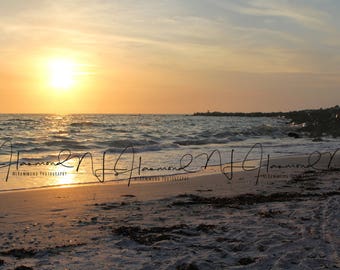 Pode incluir: Um pôr do sol pitoresco sobre uma praia de areia com ondas suaves que chegam à costa. O céu é de um laranja e rosa vibrantes, com nuvens que refletem a luz quente. O sol se põe à distância, projetando um brilho dourado sobre a água.