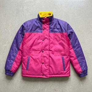 Puede incluir: Una chaqueta de bloques de color con cuerpo rosa brillante, mangas y paneles superiores en color morado, y un cuello alto forrado de forro polar amarillo. La chaqueta incluye un cierre frontal con botones a presión y dos bolsillos laterales.