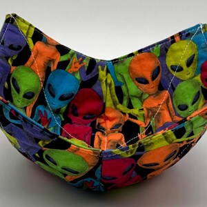 May include: A fabric bowl featuring a colorful alien print. The bowl is square-shaped with folded corners, showcasing a repeating pattern of green, blue, orange, and red aliens on a black background. White stitching outlines the edges.