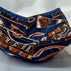 May include: A navy blue and orange fabric bowl with a Chicago Bears logo and text. The bowl is folded into a square shape with four raised corners. The fabric has a white stitched border.
