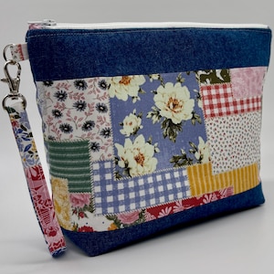 May include: A patchwork fabric pouch with a denim blue top and bottom. The pouch features a variety of floral and patterned fabrics in shades of blue, pink, red, and yellow. A matching fabric wrist strap is attached with a silver clasp.
