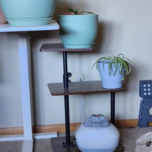 May include: A three-tiered plant stand with dark brown shelves and black metal supports. The stand holds several potted plants in various shades of green and blue. A white humidifier sits on the bottom shelf, and a white desk is visible on the left.