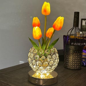 May include: A decorative lamp featuring a spherical, crystal-studded vase holding artificial orange and yellow tulips. The lamp has a gold-colored base and emits a warm glow, creating a cozy ambiance. The lamp is a decorative home accent.