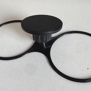 Puede incluir: Un portavasos de coche de plástico negro con una plataforma circular central y dos soportes circulares a cada lado. El portavasos está diseñado para encajar en la ranura del portavasos existente de un coche.