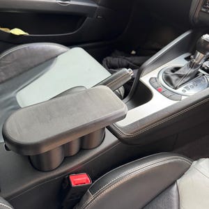May include: Black car interior with a centre console featuring a built-in armrest and cup holders. The seats are black leather with white stitching. A red seatbelt buckle is visible.