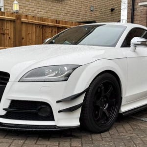 May include: A white sports car with black wheels and accents. The car has a sleek design with a low profile and a black front grill. The car is parked on a brick driveway.