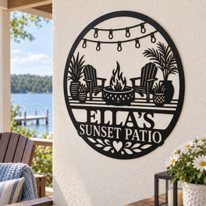 May include: A circular black metal wall sign featuring a cutout design of two chairs, a fire pit, potted plants, and string lights. The text ELLA'S SUNSET PATIO is centered in the middle, with a small heart icon at the bottom.