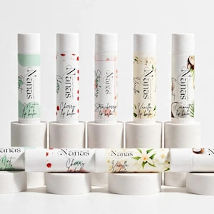 May include: A collection of nine cylindrical lip balm tubes labeled Nanas, arranged in two rows on white circular pedestals. The tubes feature various designs including mint, cherry, strawberry, vanilla, and coconut, with text identifying each flavor.