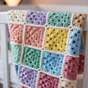 May include: A colourful, handmade crochet blanket with a granny square design. The blanket features squares in pastel shades of pink, yellow, blue, green, and purple, bordered by white yarn. The blanket is draped over a white cot.