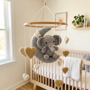 Puede incluir: Móvil para bebé hecho a mano con un elefante gris de ganchillo sobre una luna creciente gris. El móvil incluye aros de madera, corazones colgantes crema y dorados, y una estrella. Cuelga sobre una cuna de madera.