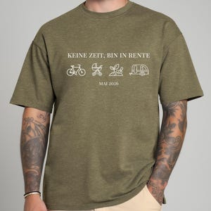 May include: Olive green t-shirt with white text and line art illustrations. The text reads "KEINE ZEIT, BIN IN RENTE" above a bicycle, stroller, plant, and camper. Below is "MAI 2026".
