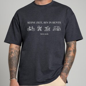 May include: Dark gray t-shirt with white text and line drawings. The text reads "KEINE ZEIT, BIN IN RENTE" above a bicycle, stroller, plant, and camper icon, with "MAI 2026" below. The shirt is a casual style.