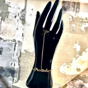 Puede incluir: Una pulsera de cadena de mano Proverbs de color dorado. La pulsera presenta un anillo que se conecta a una cadena que cae hasta la muñeca, deletreando la palabra "Proverbs". La joya se exhibe en un maniquí negro con forma de mano.