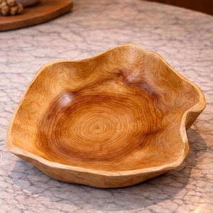 Könnte beinhalten: Eine handgefertigte Holzschale mit einer natürlichen, organischen Form. Die Schale besteht aus hellbraunem Holz mit dunkleren braunen Maserungsmustern. Das Innere weist konzentrische Kreise auf, und die Ränder haben ein unregelmäßiges, wellenförmiges Design. Diese dekorative Schale eignet sich zum Servieren oder Ausstellen.
