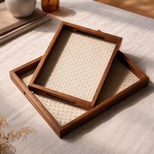 Puede incluir: Dos bandejas rectangulares de madera para servir con inserciones tejidas de color crema están apiladas sobre una superficie de color claro. Las bandejas tienen marcos de madera marrón oscuro con asas recortadas.