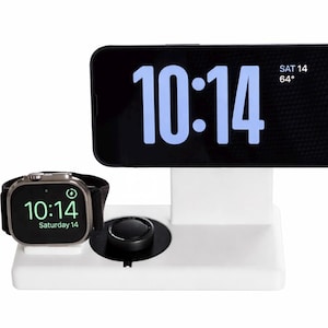 May include: A white charging station with a black phone displaying "10:14" and the date. An Apple Watch shows the same time. A black dial is centered on the base. The time and date are displayed in white.