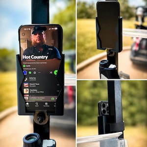 May include: Black phone holder mounted on a golf cart. A smartphone is displayed in the holder, showing a music app with the text "Hot Country" and a photo of a country music artist. The holder is attached to a black metal bar.