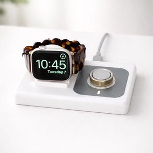 May include: A gold and white smartwatch with a tortoiseshell band rests on a white charging dock. The watch face displays the time 10:45 and the date Tuesday 7. A silver and gold charging puck is also on the dock.