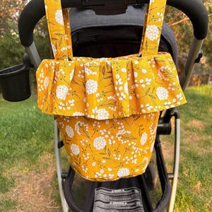 May include: A mustard yellow fabric stroller organizer bag featuring a white floral and botanical print. The bag has two top straps for hanging and a wide, pleated ruffle detail along the upper edge, providing storage space for baby essentials.