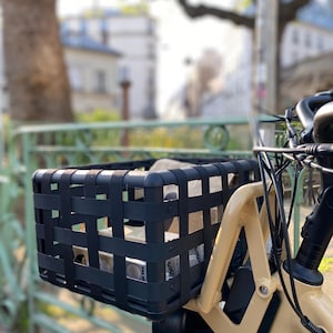 May include: A black woven bicycle basket attached to a cream-colored bicycle. The basket is rectangular and contains items. The background shows a blurred street scene with trees and buildings.
