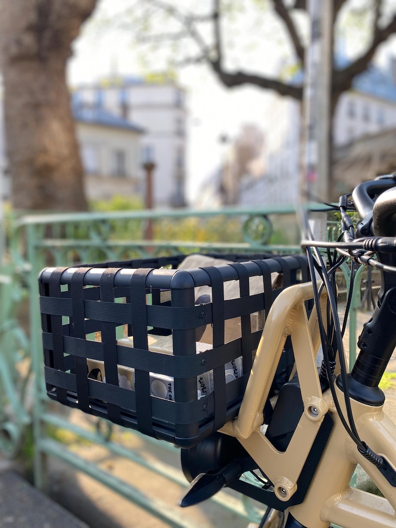 May include: A black woven bicycle basket attached to a cream-colored bicycle. The basket is rectangular and contains items. The background shows a blurred street scene with trees and buildings.