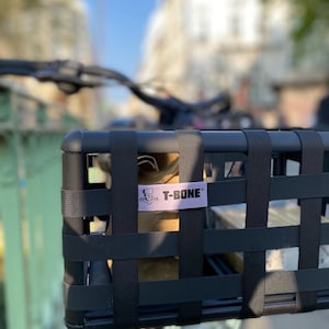 May include: Black bicycle basket with a woven design, mounted on the handlebars. The basket has a rectangular shape and features a label that reads "T-BONE" in white text. The background shows a blurred street scene with buildings and a blue sky.