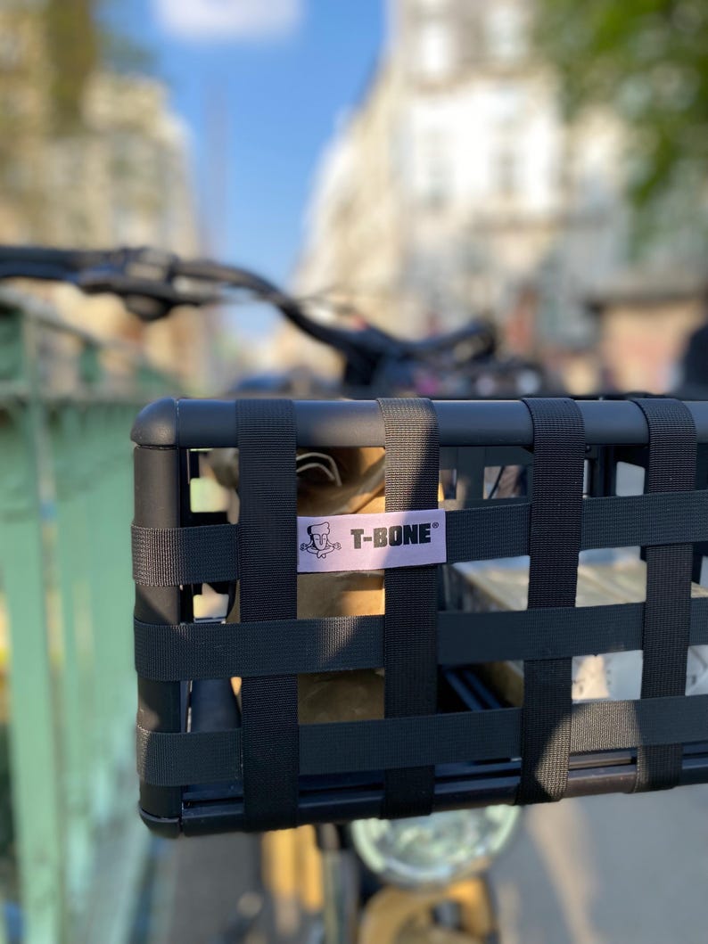 May include: Black bicycle basket with a woven design, mounted on the handlebars. The basket has a rectangular shape and features a label that reads "T-BONE" in white text. The background shows a blurred street scene with buildings and a blue sky.