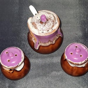 Peut inclure: Un ensemble de condiments en céramique comprenant un sucrier avec une cuillère et deux salières. L'ensemble a une base marron avec un glaçage violet et blanc. Le sucrier a un couvercle avec un bouton violet. Les salières ont trois trous sur le dessus.