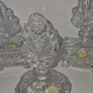 May include: Clear crystal perfume bottles with ornate leaf-shaped stoppers. The bottles have a detailed, textured design and are arranged on a white surface. The image showcases the intricate craftsmanship of the glasswork.