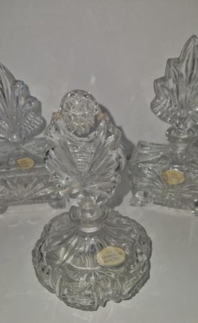 May include: Clear crystal perfume bottles with ornate leaf-shaped stoppers. The bottles have a detailed, textured design and are arranged on a white surface. The image showcases the intricate craftsmanship of the glasswork.