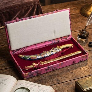 May include: An open pink floral box with a decorative gold and blue knife and pen set. The set includes a gold-colored pen and a knife with a blue and gold design. The set is displayed on a wooden table with a ledger and magnifying glass.