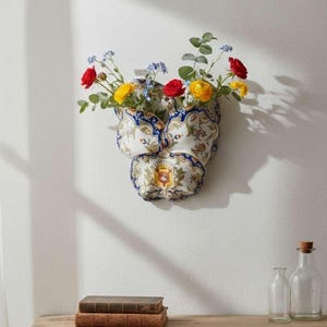 Könnte beinhalten: Eine Wandvase aus Keramik mit floralem Design, gefüllt mit roten, gelben und blauen Blumen. Die Vase hat ein blau-weißes Muster mit einem Wappen in der Mitte. Zwei alte Bücher und Glasflaschen stehen auf einem Holztisch.