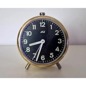 May include: A vintage brass-colored alarm clock with a black face. The clock features white numerals and hands, with the brand name "JAZ" printed at the top. The clock has a round shape and stands on two small legs.
