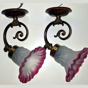 Peut inclure: Une paire d'appliques murales dotées de plaques de montage circulaires marron foncé, de bras métalliques incurvés couleur bronze avec des volutes, et d'abat-jours en verre dépoli évasés aux bords ondulés teintés de rose foncé.