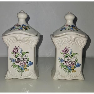 Può includere: Due barattoli in ceramica color bianco sporco con coperchi abbinati, ciascuno decorato con un bouquet floreale e un nastro con la scritta Poivre o Sel. I contenitori hanno base quadrata, motivo a griglia in rilievo e un pomello.