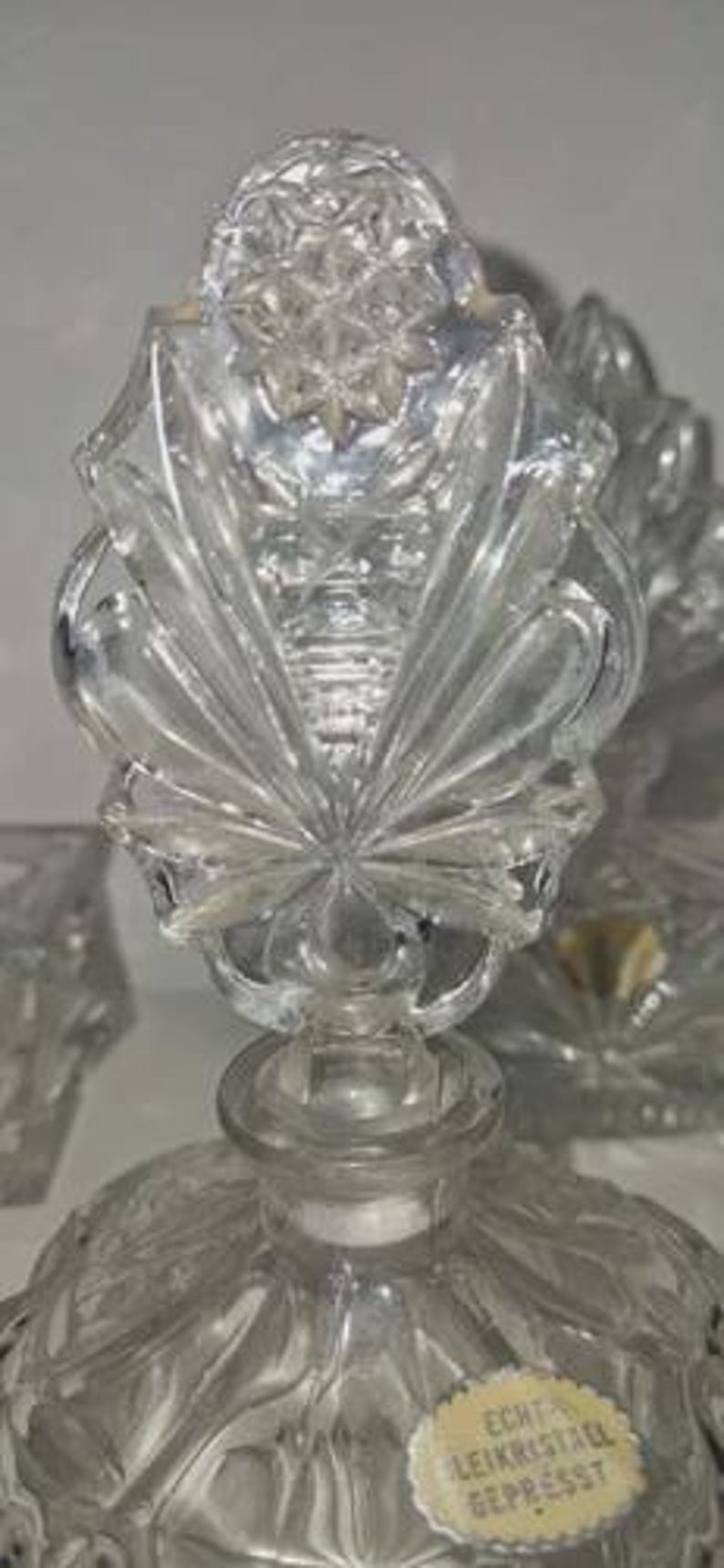 May include: Clear crystal perfume bottle with a decorative stopper. The stopper features a starburst design with a detailed, textured pattern. The bottle has a round base and a label that reads "Echt Leikristall Gepresst".