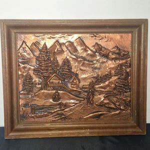 May include: A framed copper relief art piece depicting a mountain landscape with cabins, trees, and figures. The artwork is in a brown wooden frame. The relief is in shades of copper and brown, with detailed textures of the mountains and trees.