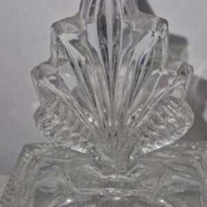 May include: Clear crystal glass decorative box with a leaf-shaped lid. The box has a rectangular base with a detailed, textured design. The lid features a fan-like pattern. A small, gold-colored sticker is affixed to the front.