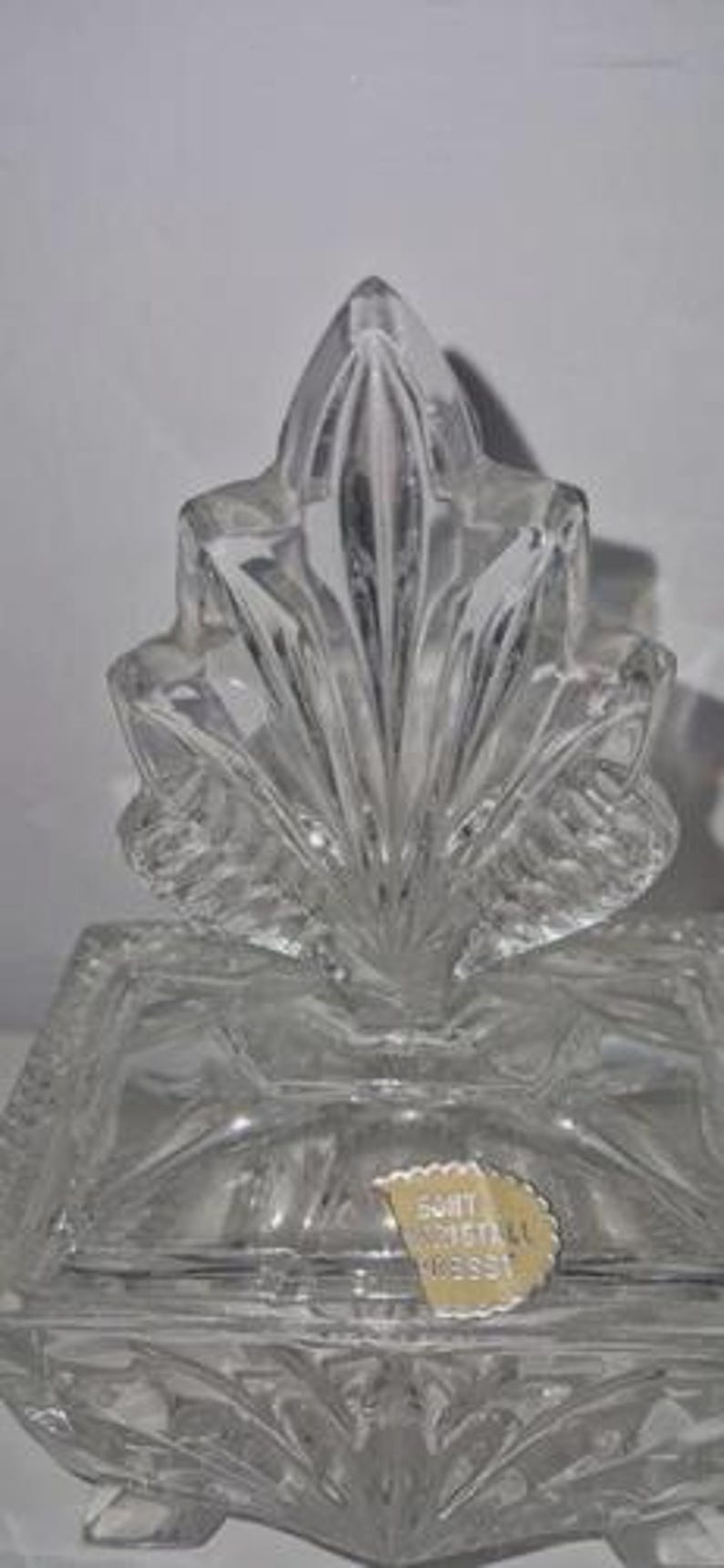 May include: Clear crystal glass decorative box with a leaf-shaped lid. The box has a rectangular base with a detailed, textured design. The lid features a fan-like pattern. A small, gold-colored sticker is affixed to the front.