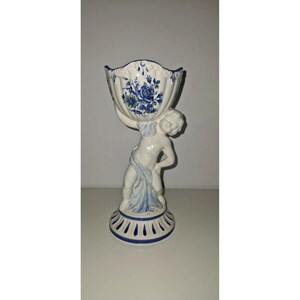 Peut inclure: Un vase en céramique blanche représentant une figurine sculptée tenant une coupe festonnée. La coupe est ornée de motifs floraux bleus et la base présente un liseré bleu. La figurine est drapée d'un tissu bleu clair, avec une finition brillante.