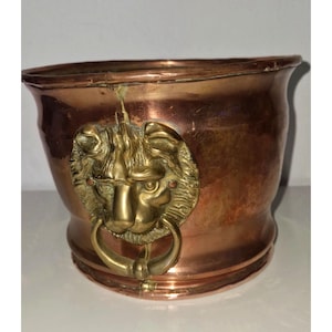 May include: A copper-colored metal planter with a brass lion's head door knocker. The planter has a rounded shape with a slightly flared rim. The lion's head is detailed with a ring for a handle.