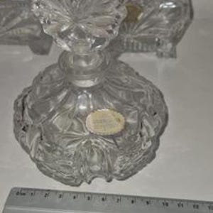 May include: A clear, cut-glass perfume bottle with a decorative stopper. The bottle has a round base and a detailed, textured design. A ruler is placed near the bottle, providing a size reference. The bottle is likely vintage or antique.
