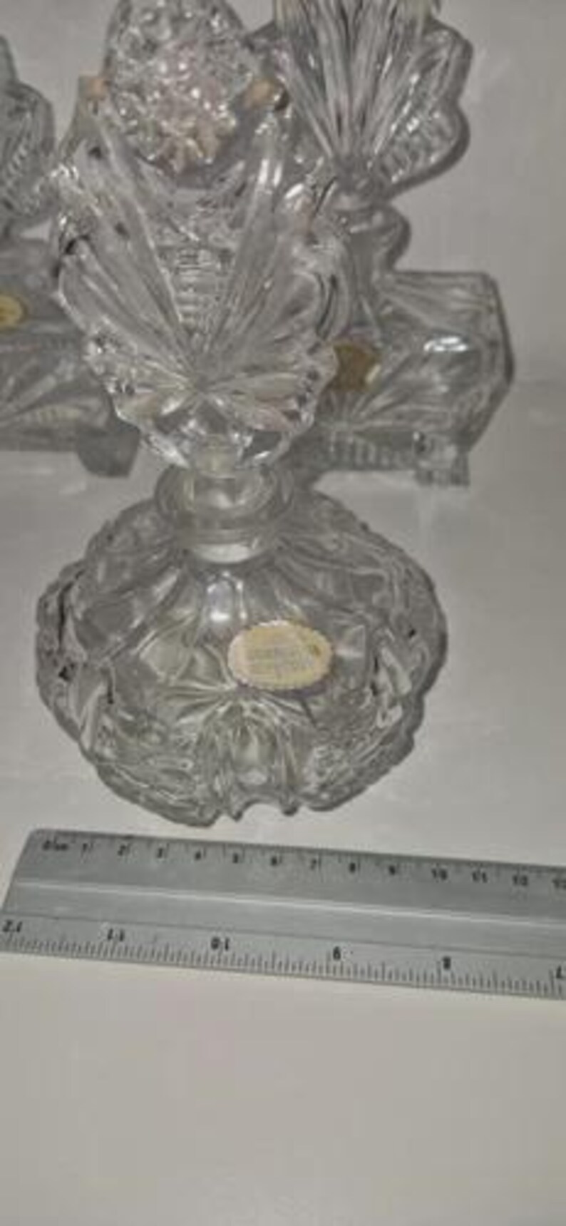 May include: A clear, cut-glass perfume bottle with a decorative stopper. The bottle has a round base and a detailed, textured design. A ruler is placed near the bottle, providing a size reference. The bottle is likely vintage or antique.