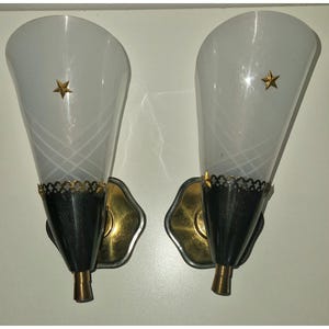 Op de afbeelding: Een paar wandlampen met conische, matglazen kappen voorzien van witte kruispatronen en gouden steraccenten. De basis van elke kap heeft een zwarte band met een decoratieve gouden schulprand, gemonteerd op een goudkleurige metalen muurplaat.