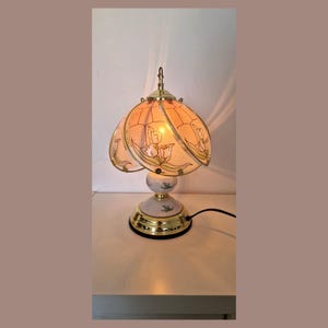 Peut inclure: Une lampe de table vintage avec un motif floral. La lampe présente un abat-jour pêche avec un motif de tulipes, une base dorée et un corps en céramique blanche. La lampe est allumée, projetant une lueur chaleureuse.