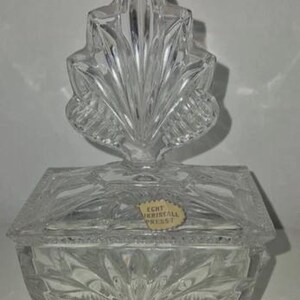 May include: A clear, rectangular crystal box with a leaf-shaped finial. The box has a lid and sits on four small feet. The crystal is intricately cut with a sunburst pattern on the front and sides. The box has a sticker that reads "ECHT KRISTALL PRESS?"