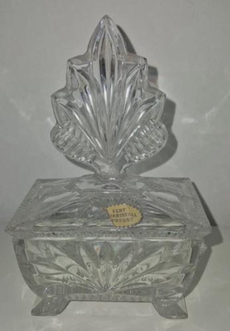 May include: A clear, rectangular crystal box with a leaf-shaped finial. The box has a lid and sits on four small feet. The crystal is intricately cut with a sunburst pattern on the front and sides. The box has a sticker that reads "ECHT KRISTALL PRESS?"
