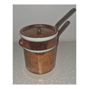 May include: A cylindrical copper double boiler with a hammered texture on the lower section. It features a white ceramic rim, a matching copper lid with a small central knob, and two long, angled metal handles extending from the side.