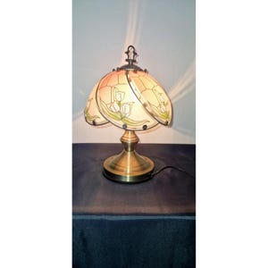 Peut inclure: Une lampe de table en laiton avec un abat-jour en vitrail. L'abat-jour présente un motif floral avec des fleurs blanches et des feuilles vertes. La lampe a une base ronde et une tige incurvée. La lampe est branchée sur une prise électrique.