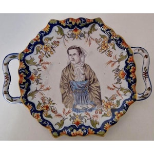Peut inclure: Un plateau de service décoratif en céramique avec un bord festonné et deux poignées. Le plateau présente un portrait central d'une personne en tenue traditionnelle, entouré de motifs floraux et foliacés bleus, jaunes et oranges.
